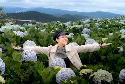 Photo of Smiling Woman in Black Hat, White Coat, and Glasses Standing in Middle of Hydrangea Flower Field With Her Eyes Closed and Hands Stretched Out