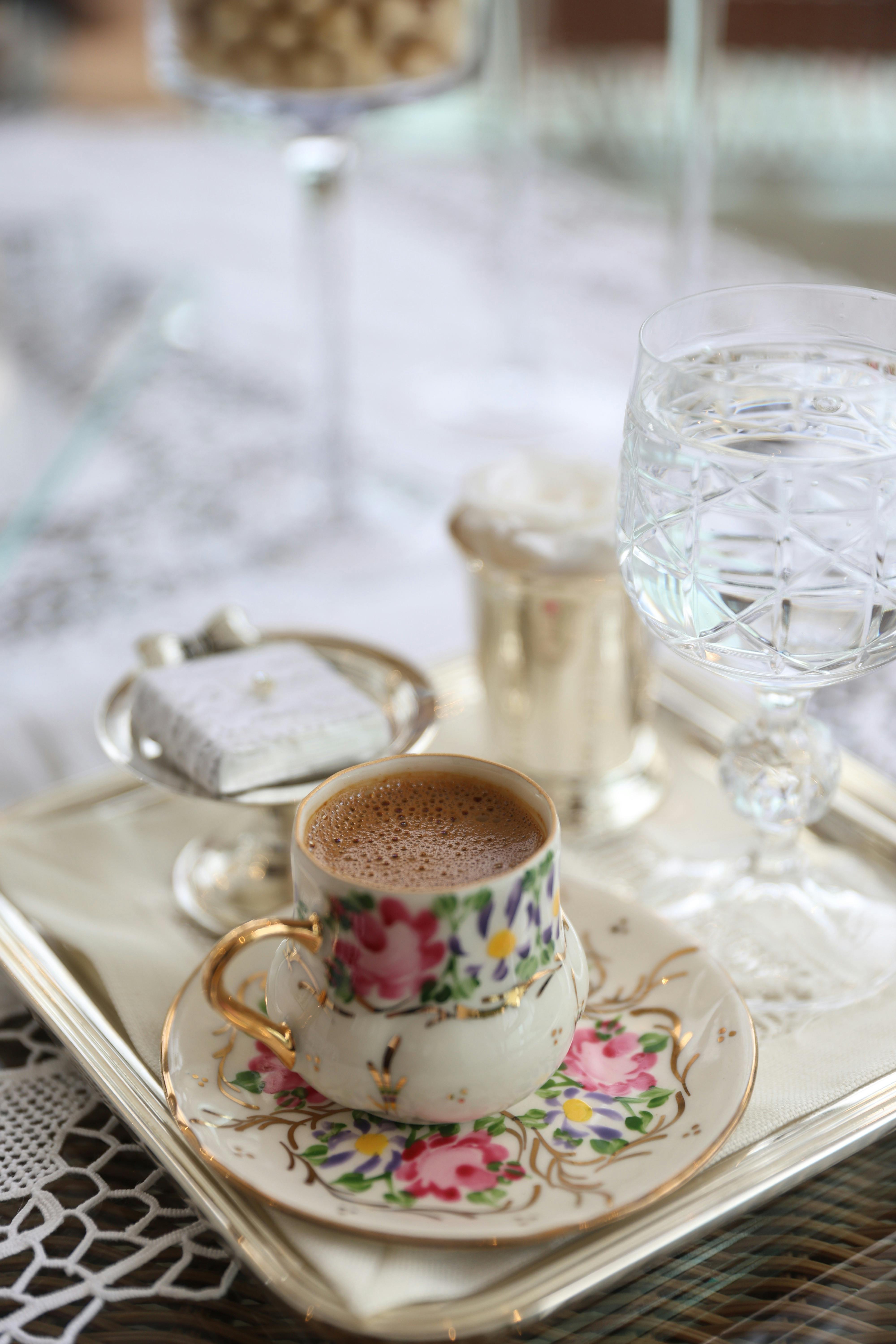A close-up of a floral teacup with coffee, a glass, and a decorative tray.