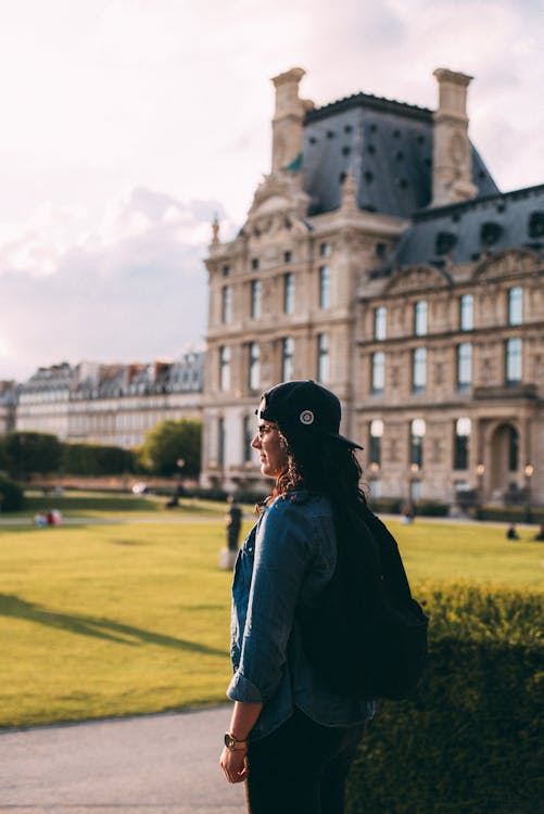 Free Vue Latérale Photo De Femme Debout En Casquette Noire Et Chemise En Jean Bleu Sac à Dos Avec Bâtiment En Arrière Plan Stock Photo