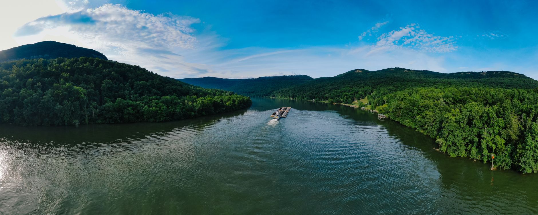 Aerial panorama of a barge navigating a lush, green river surrounded by mountains.