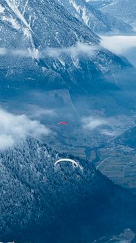 Paragliders soar through misty skies over snow-covered mountains and a distant valley below.