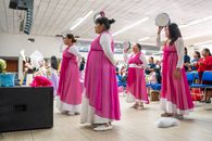Women in pink dresses performing a cultural dance with tambourines indoors.