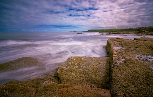 Free Serene long exposure capture of rocky shore meeting ocean waves under a dramatic sky. Stock Photo