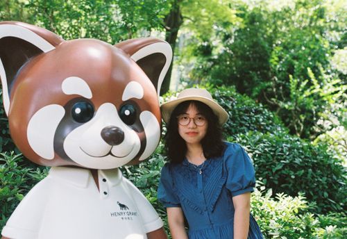 Free A young woman with glasses and a hat poses next to a red panda mascot outdoors in a lush garden. Stock Photo