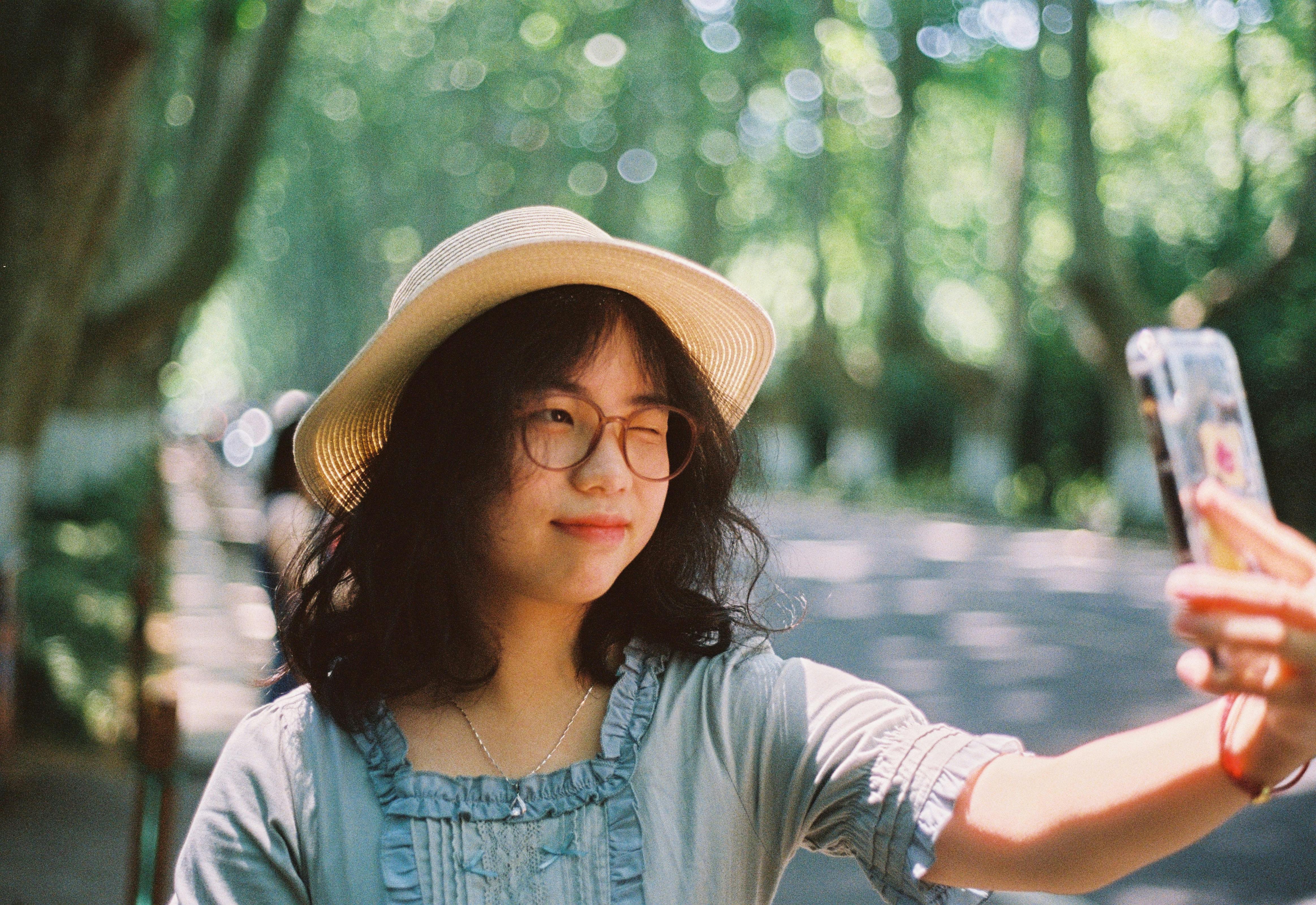Young woman with sunglasses and hat taking a selfie on a sunny day outdoors.