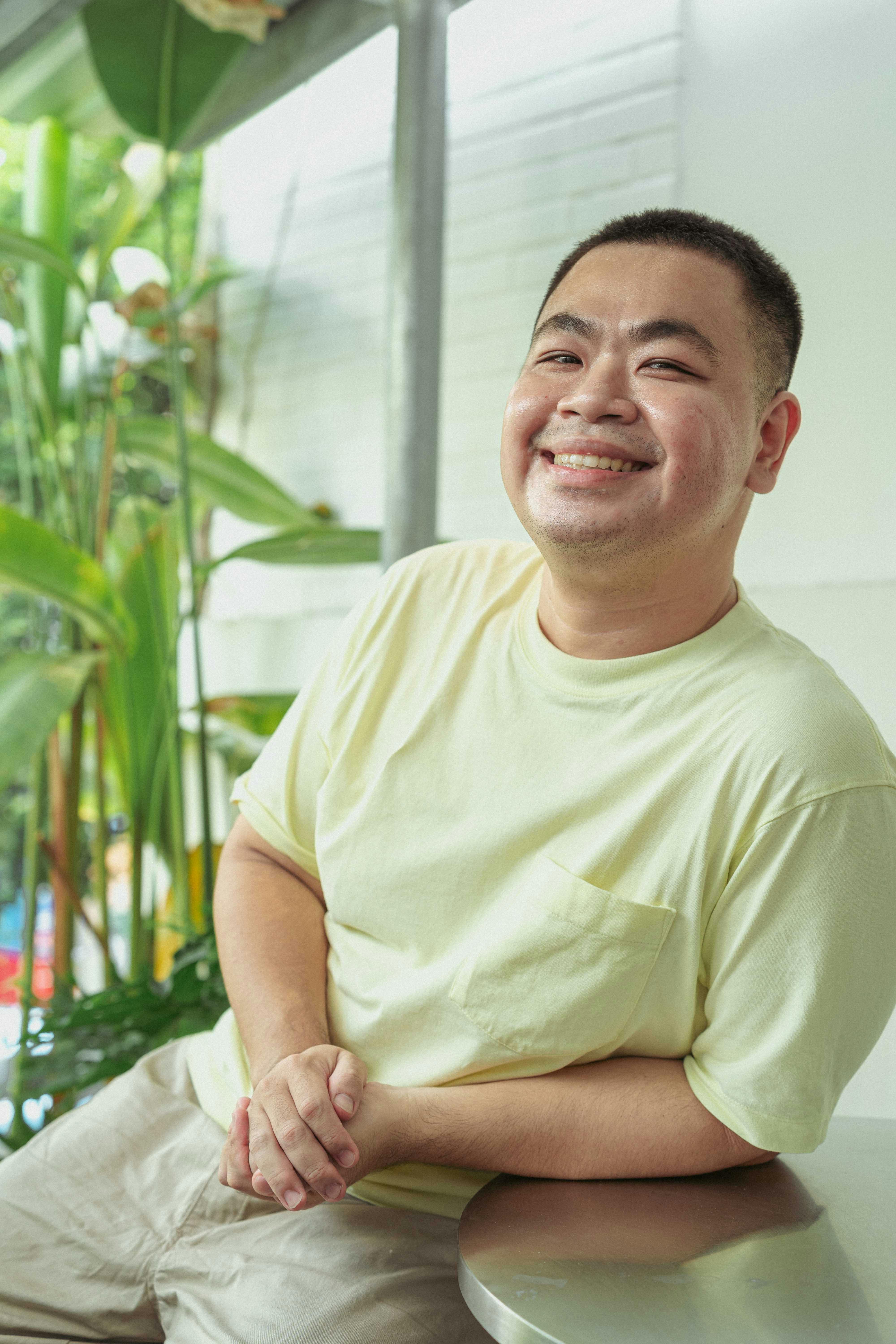 Asian man in a light t-shirt smiling indoors with plants around.