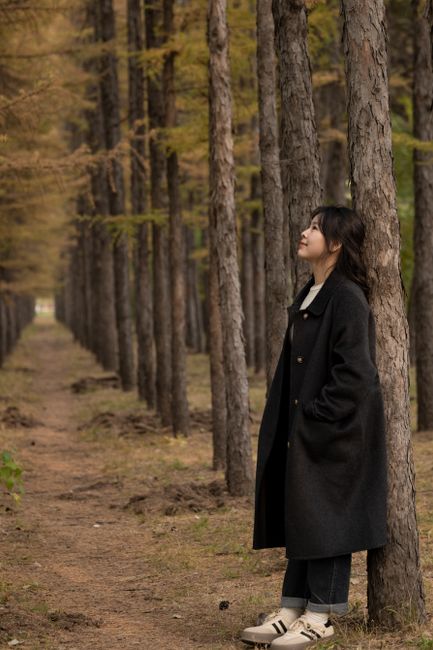 A woman in a black coat leans against a tree in a tranquil forest setting.