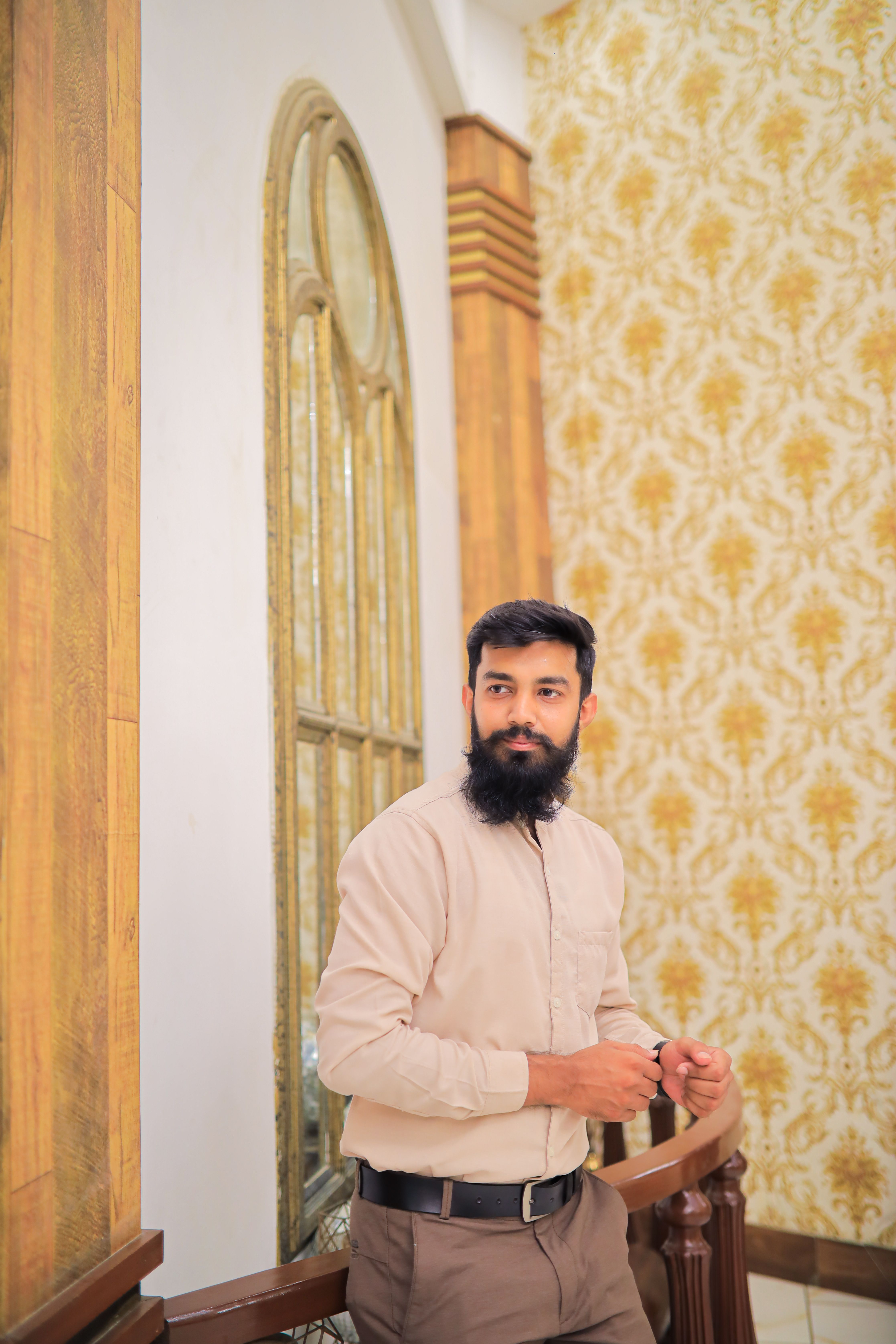 Bearded man in smart casual attire standing by ornate mirror indoors.