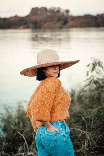 Stylish woman in denim shorts and a straw hat poses by the lake, showcasing casual fashion.