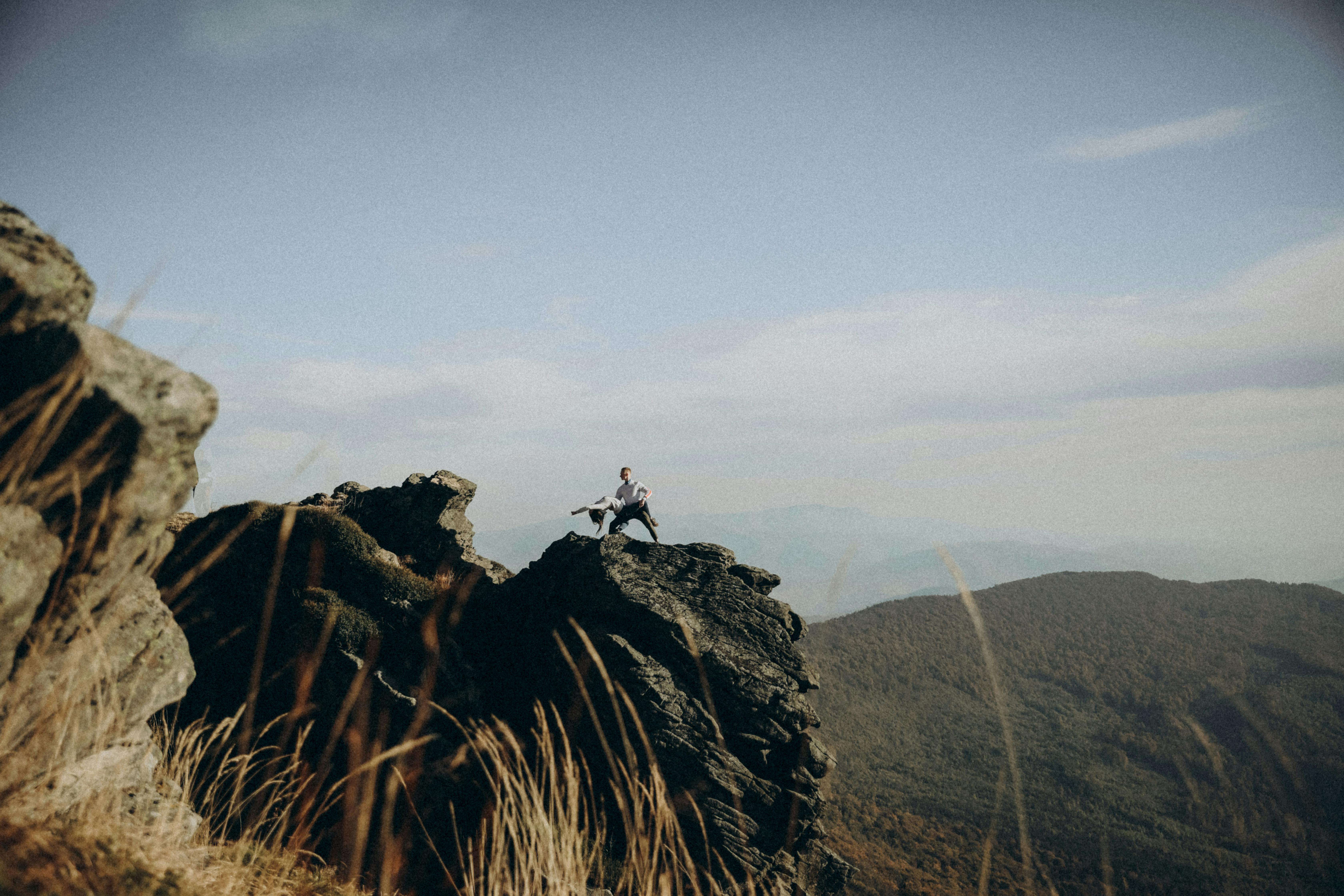 A couple gracefully dances on a rocky mountain peak under a clear sky, capturing a moment of freedom.