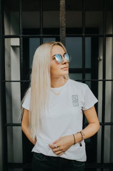 一位时尚的金发女子身着休闲装，戴着墨镜，长发飘逸地站在背景前