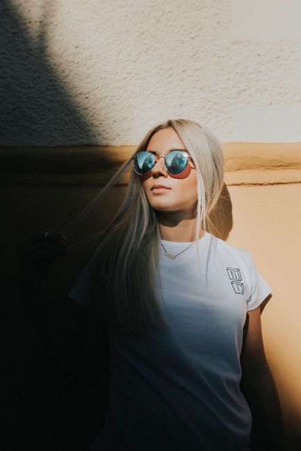 Stylish woman with long blonde hair and sunglasses posing against a textured wall in sunlight.