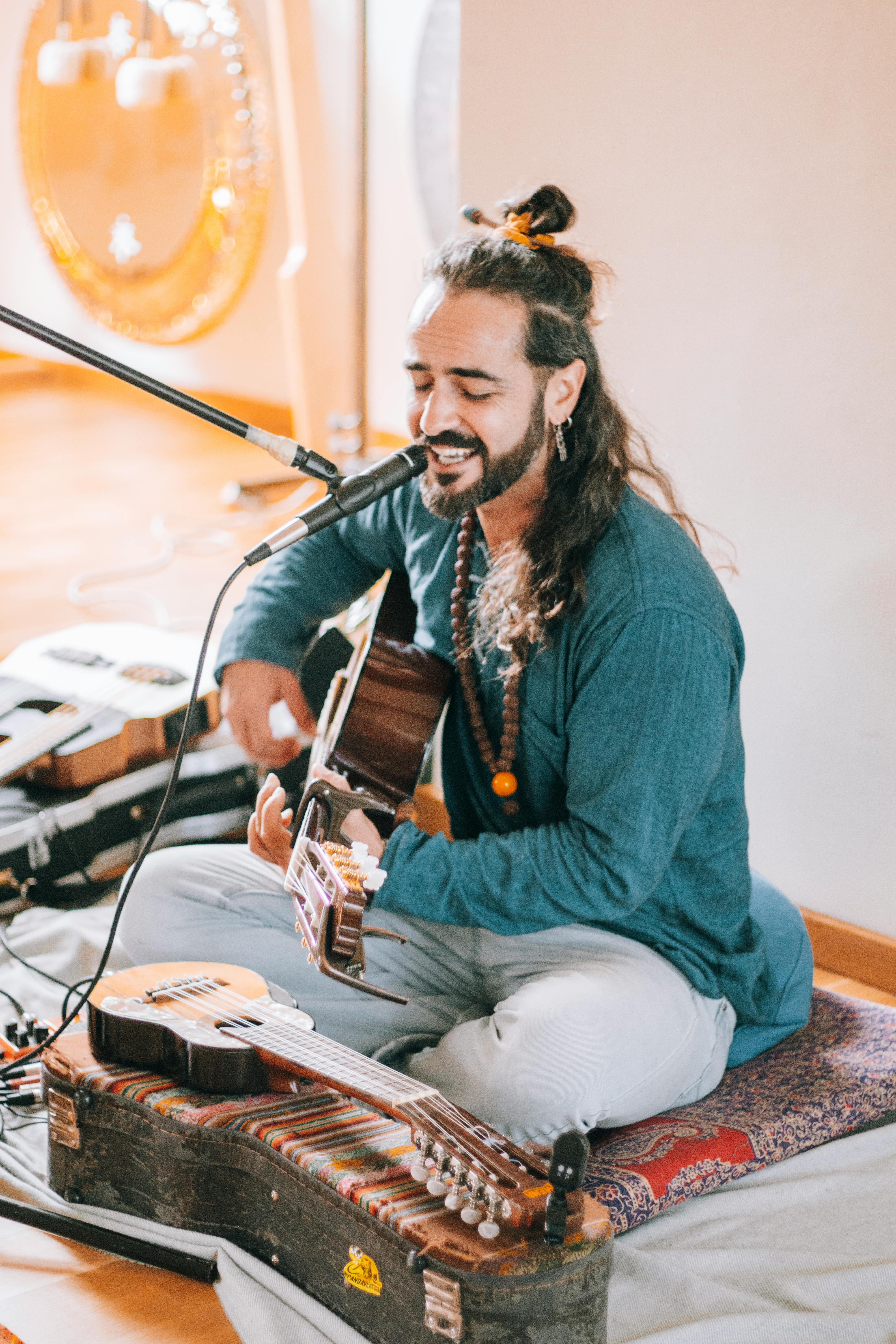 A musician with long hair performs indoors, playing the guitar and singing into a microphone.