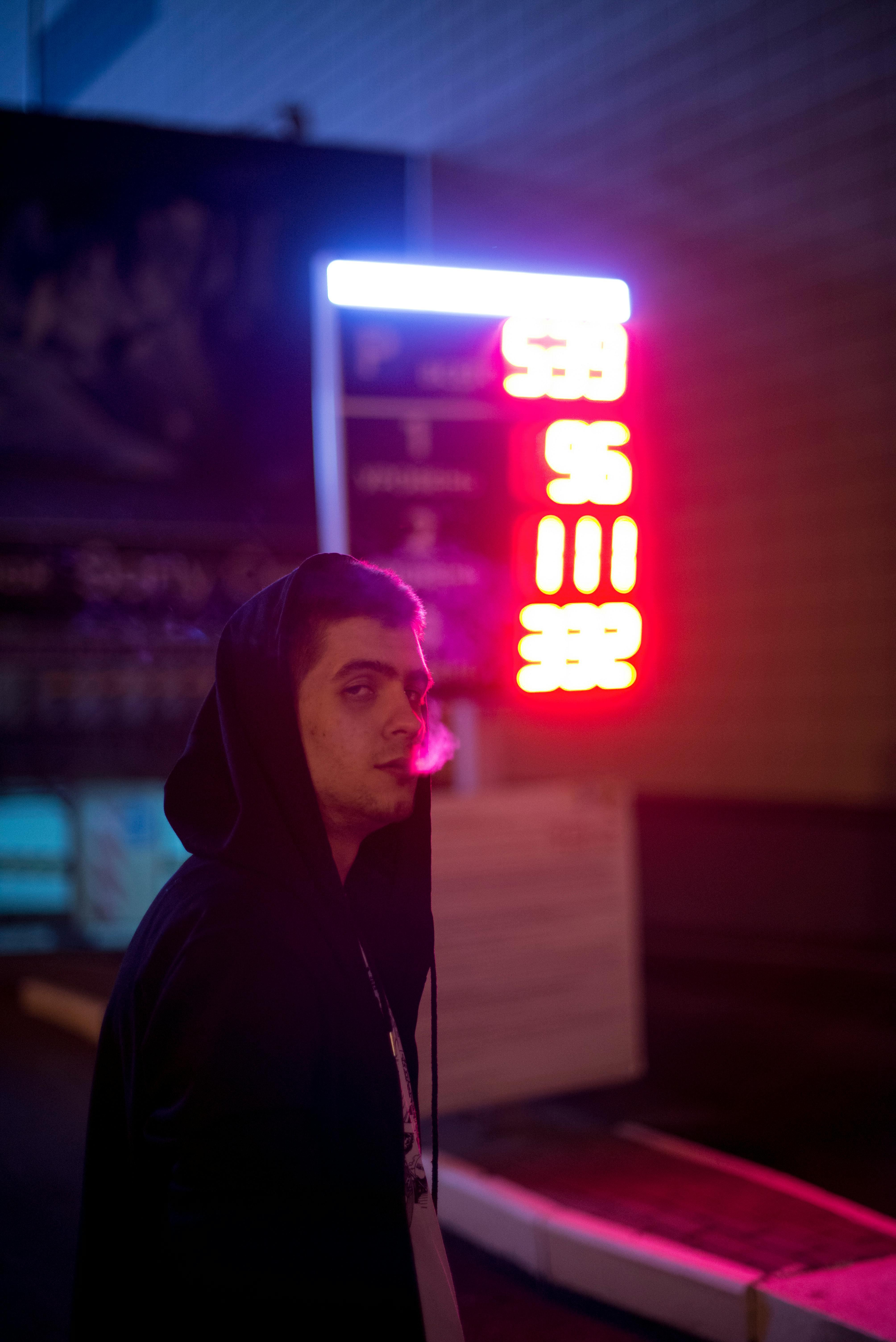 夜晚，一名穿着连帽衫的男子站在霓虹灯下，吐着烟