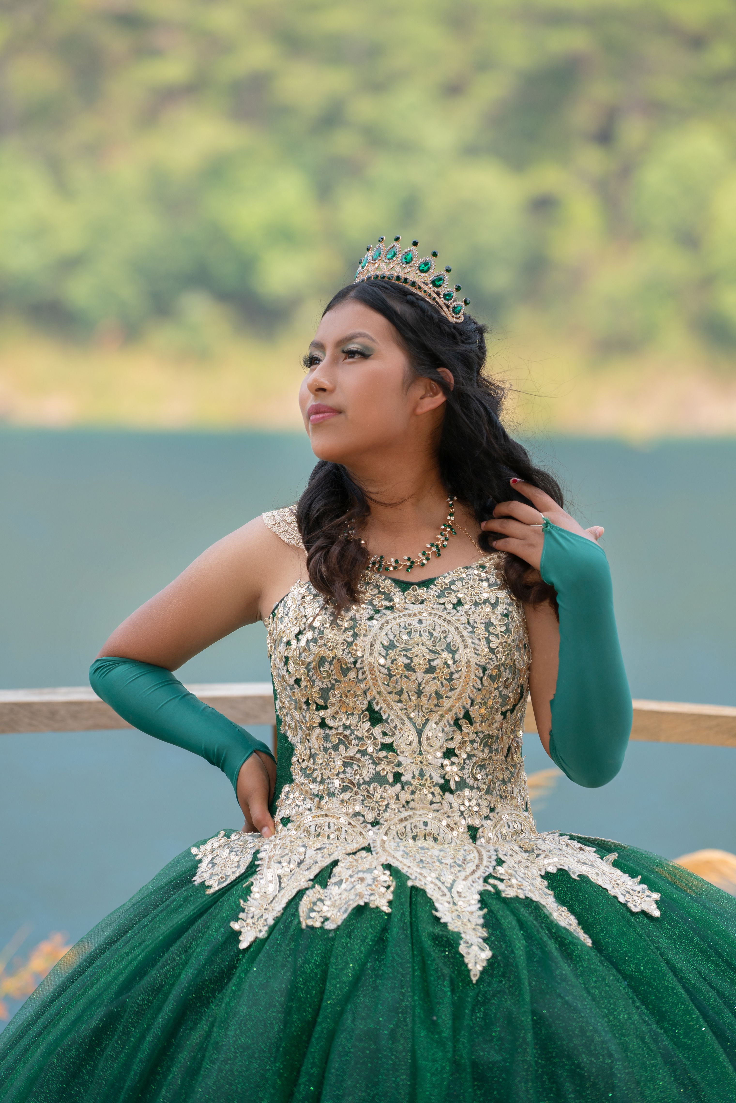 A woman in an ornate green dress and crown poses outdoors with a forest background.