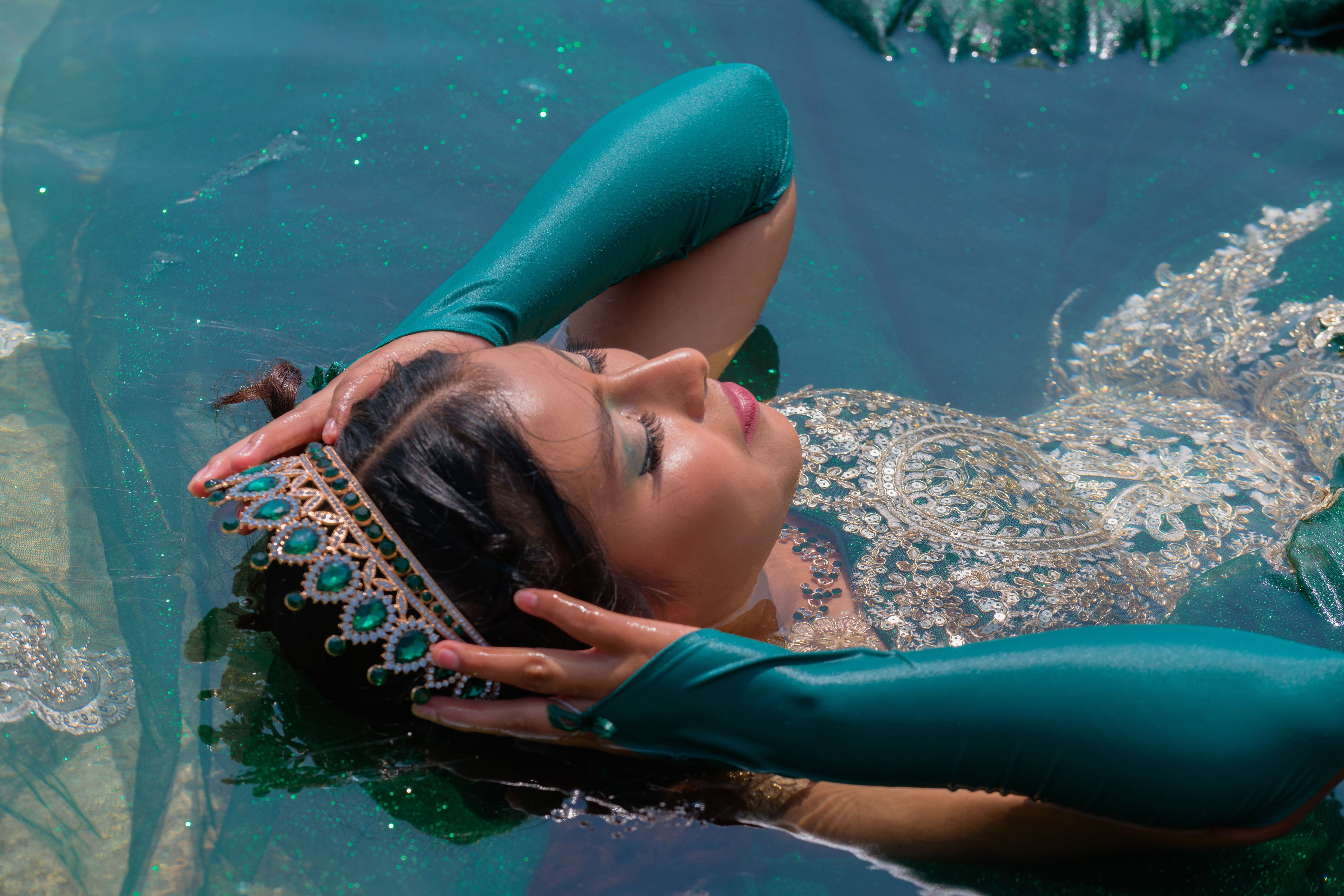 A graceful woman in a turquoise gown and jeweled crown rests serenely in shimmering water.