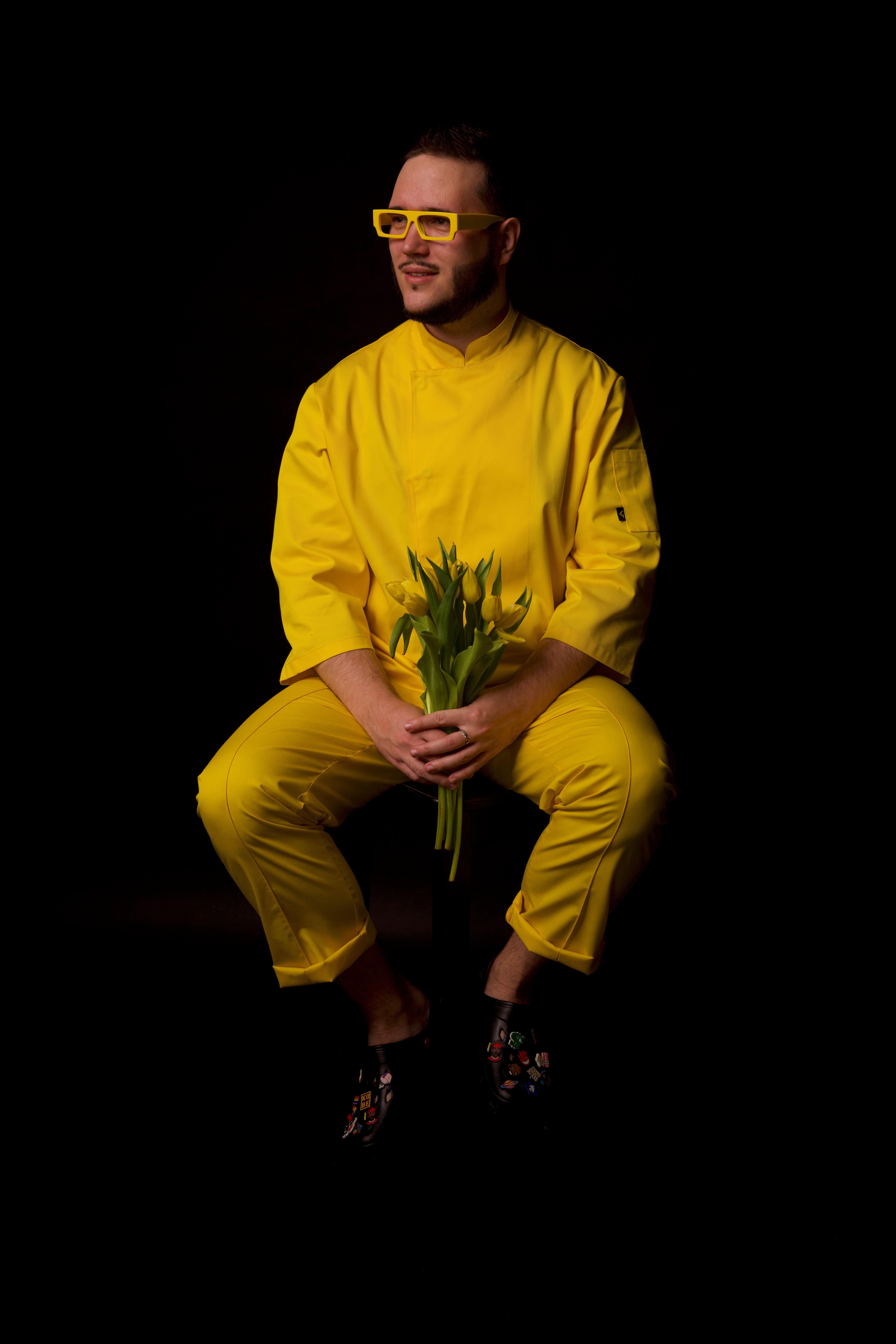 Stylish man wearing a bright yellow outfit and glasses, holding a bouquet of tulips against a dark studio background.