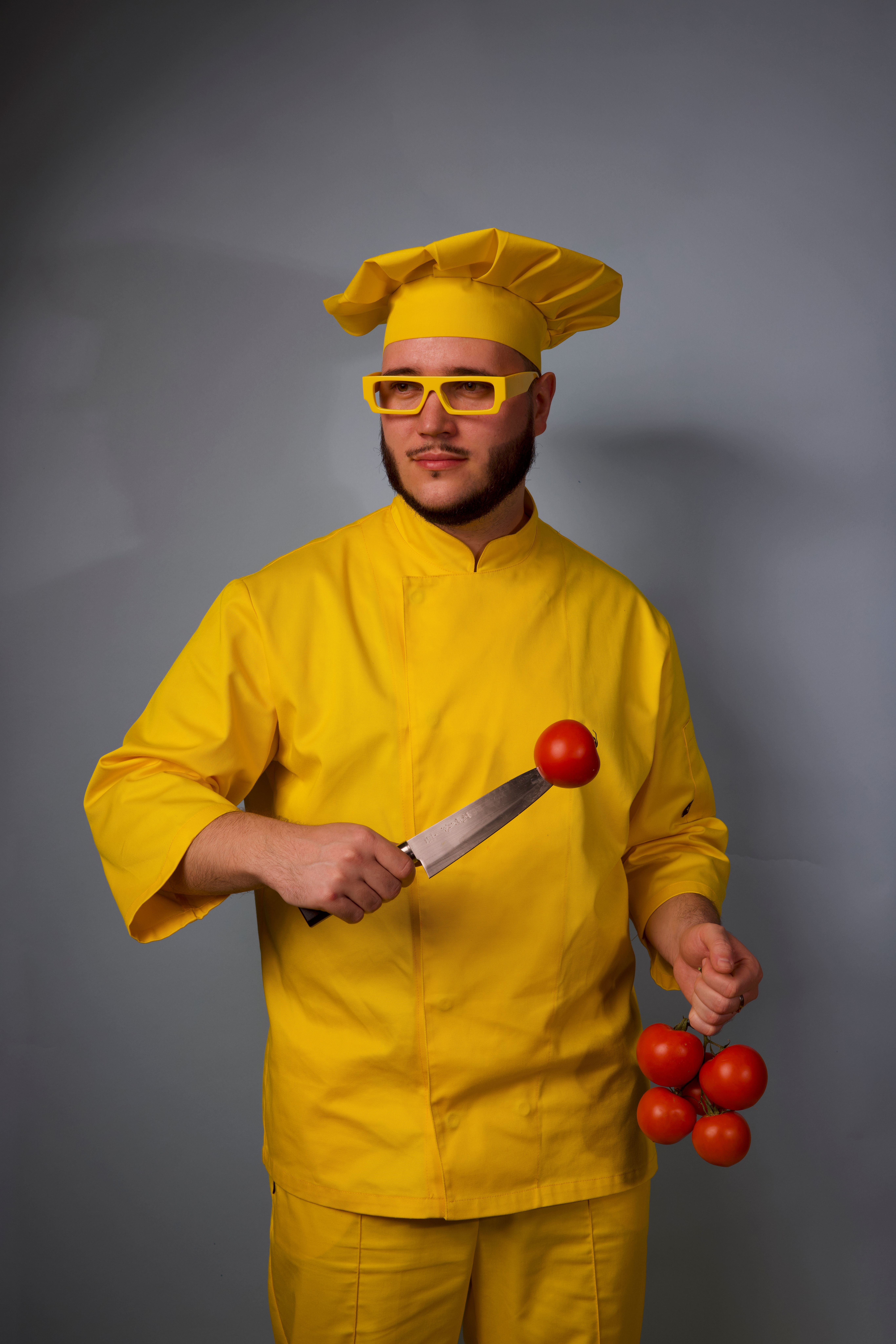 Chef in yellow outfit posing with tomatoes and knife against gray background.