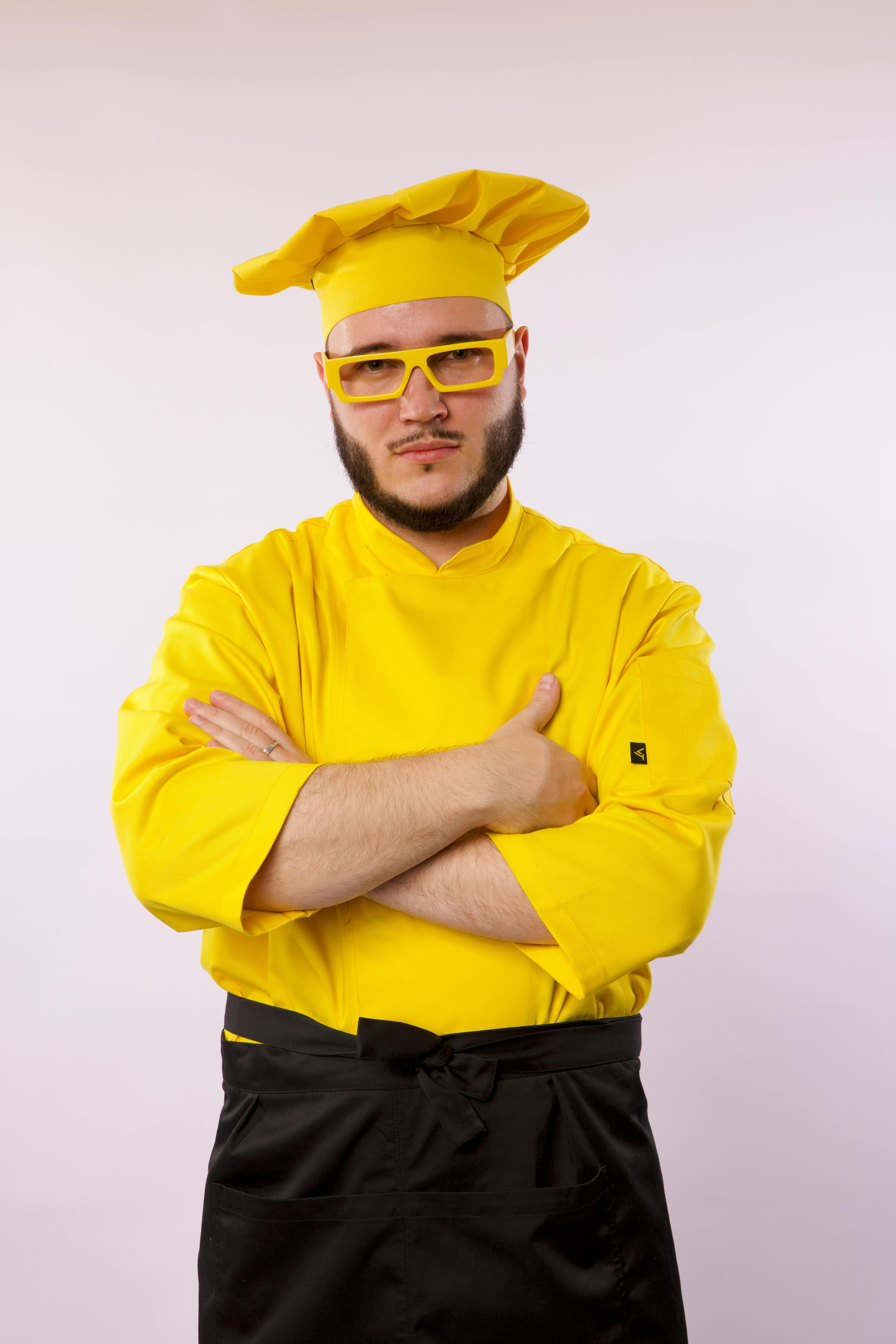 Portrait of a confident male chef in yellow attire with crossed arms and glasses against a studio background.