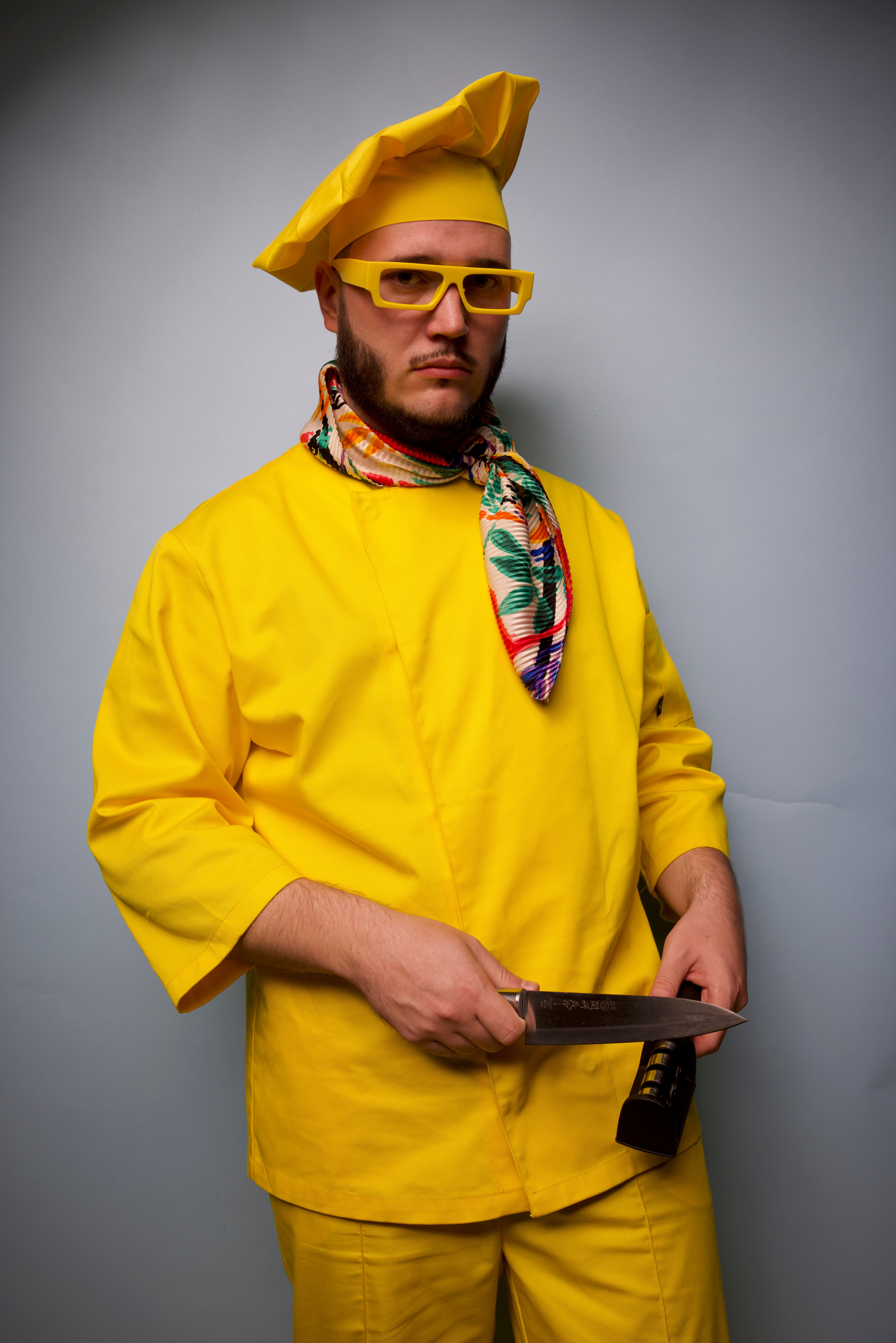 Portrait of a chef in yellow attire holding a knife with a serious expression.