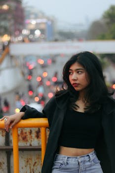 Stylish woman in black top and jacket leaning on railing in cityscape with bokeh lights.