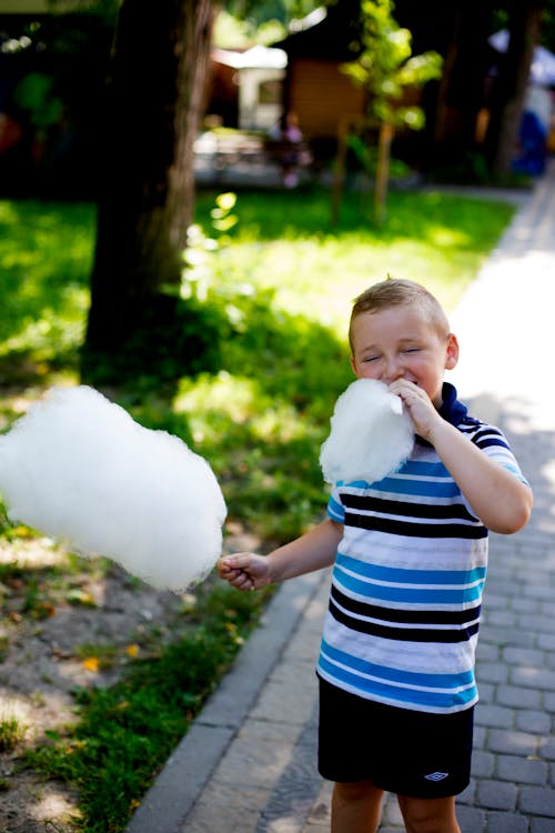 Selective Focus Photo of Boy Eating Cotton Candy with His Eyes Closed Standing on Side Walk