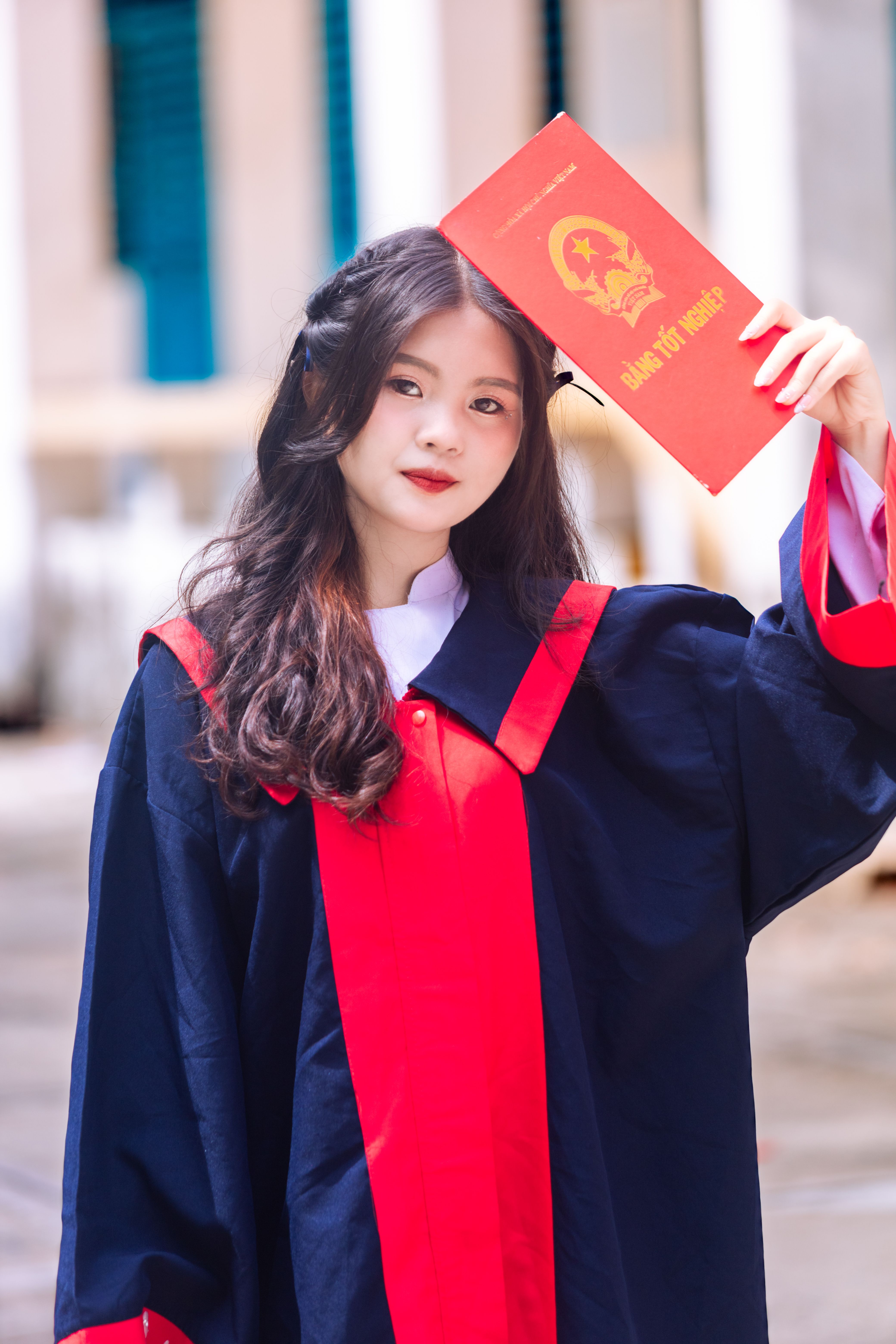 Portrait of a young Asian woman in graduation attire holding a diploma outdoors.