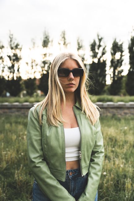 Young woman in sunglasses and green jacket standing in a field during daytime.