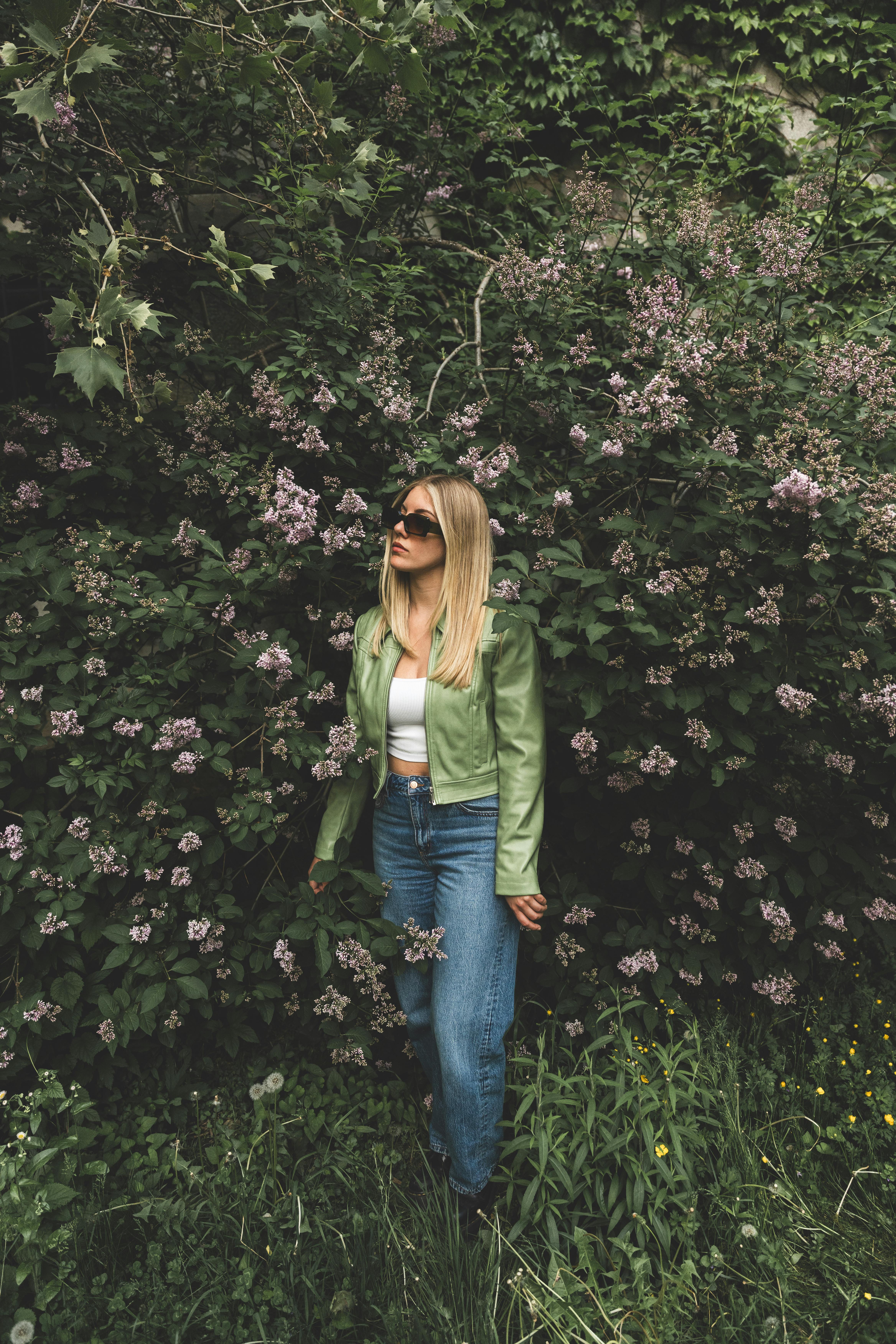 Stylish woman in green jacket posing in a lush garden with pink flowers in Montréal, Canada.