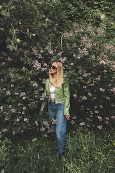 Stylish woman in green jacket posing in a lush garden with pink flowers in Montréal, Canada.