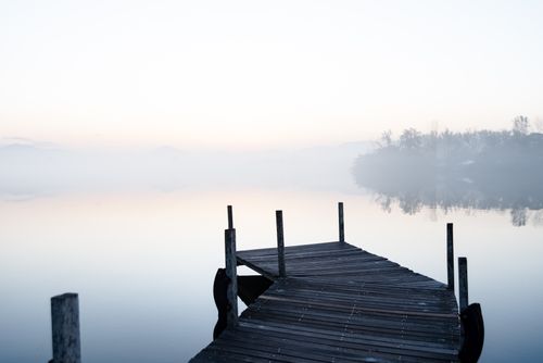 Free A tranquil lakeside view at dawn with mist enveloping the boardwalk and distant trees. Stock Photo