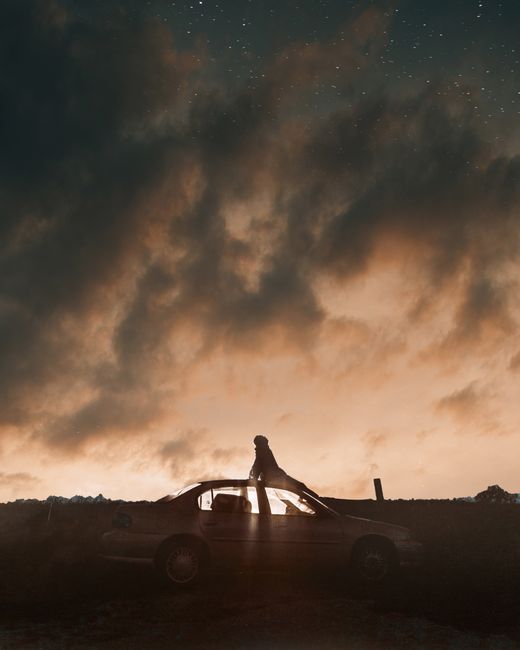 A person stands on a car roof gazing at stars against a dramatic dusk sky.