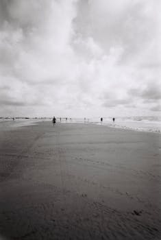 A serene black and white photo capturing people walking along a vast beach under a cloudy sky.