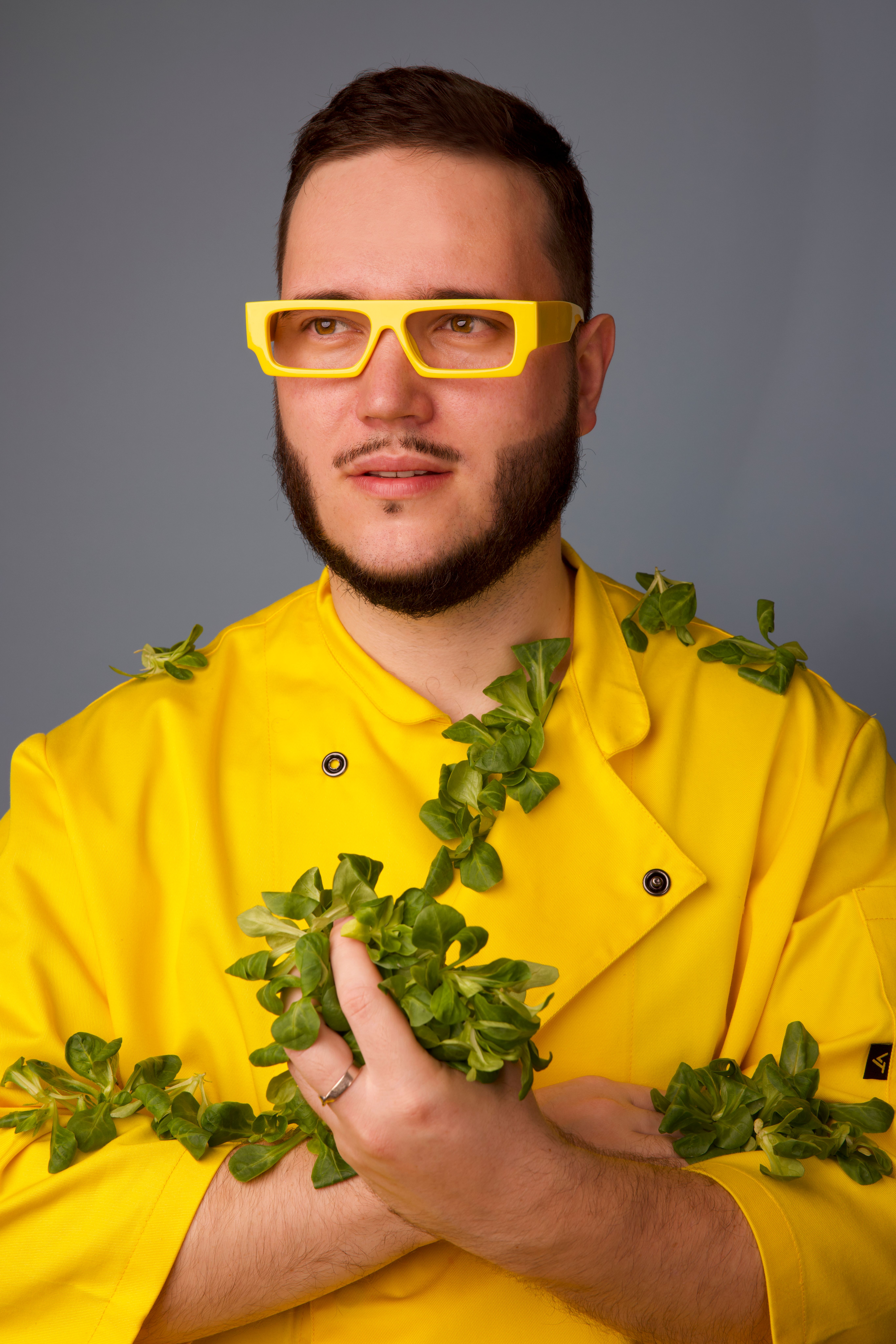 Portrait of a modern chef in vibrant yellow attire and glasses holding fresh greenery in a studio setting.