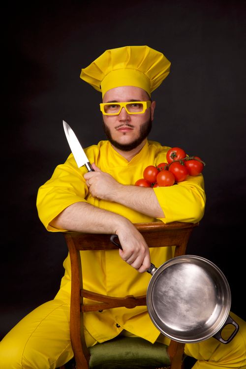 Free Chef in vibrant yellow attire holding a knife, pan, and tomatoes against a black background. Stock Photo