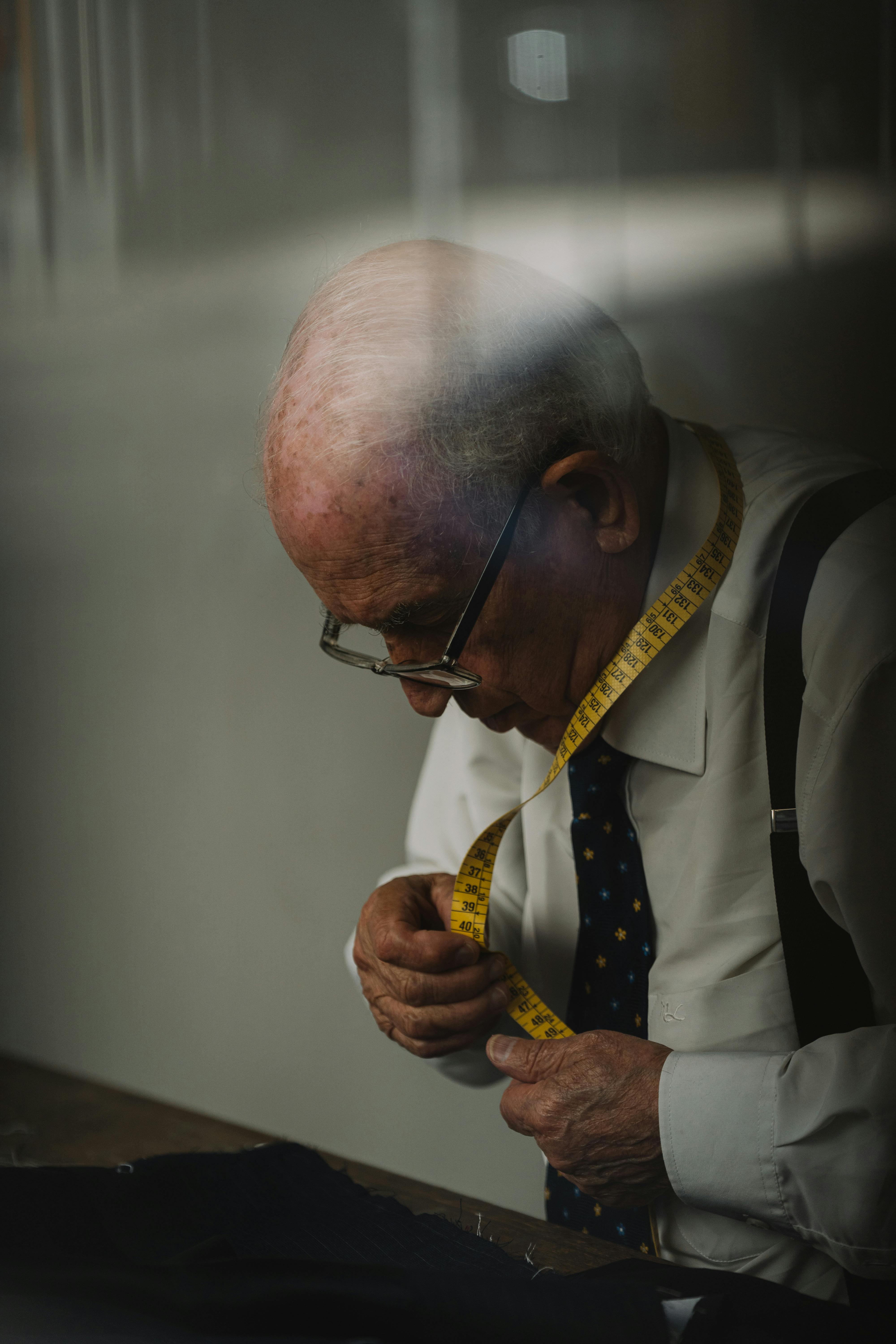 Senior man in glasses using tape measure indoors, wearing a tie and white shirt.
