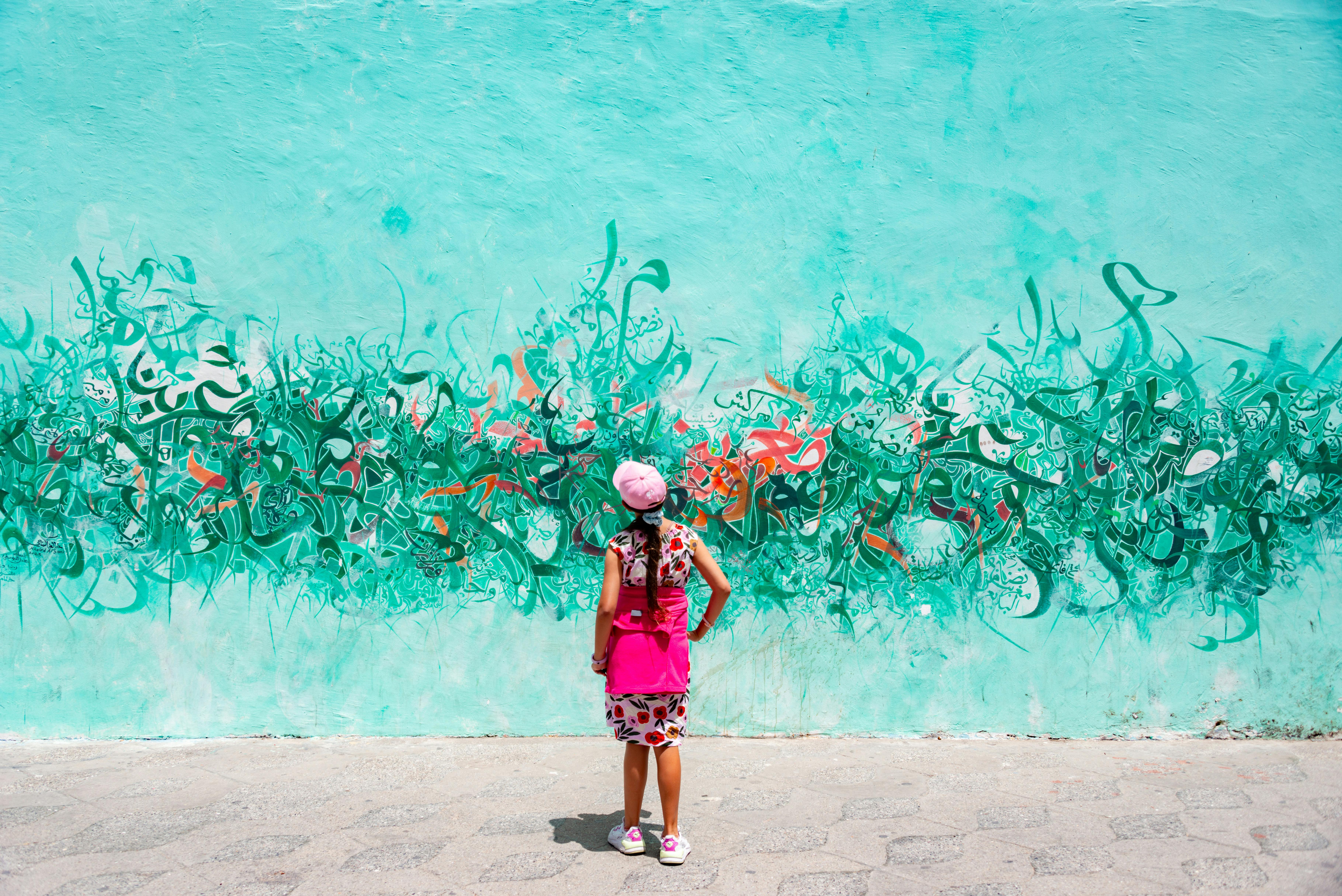 A woman in a pink outfit stands before vivid graffiti on a turquoise wall, blending urban art with fashion.