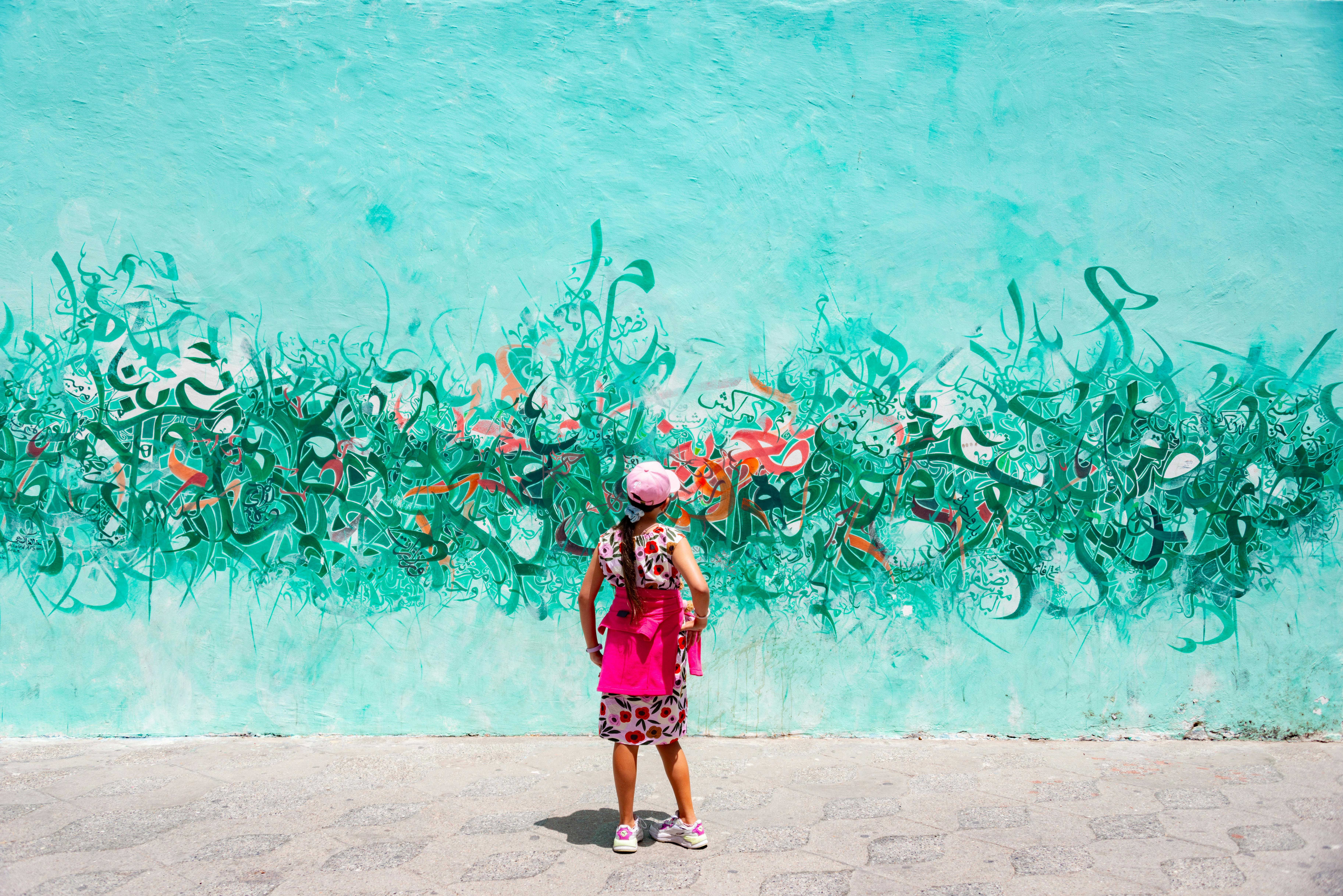 A girl admires vibrant calligraphy street art on a teal wall in an urban setting.