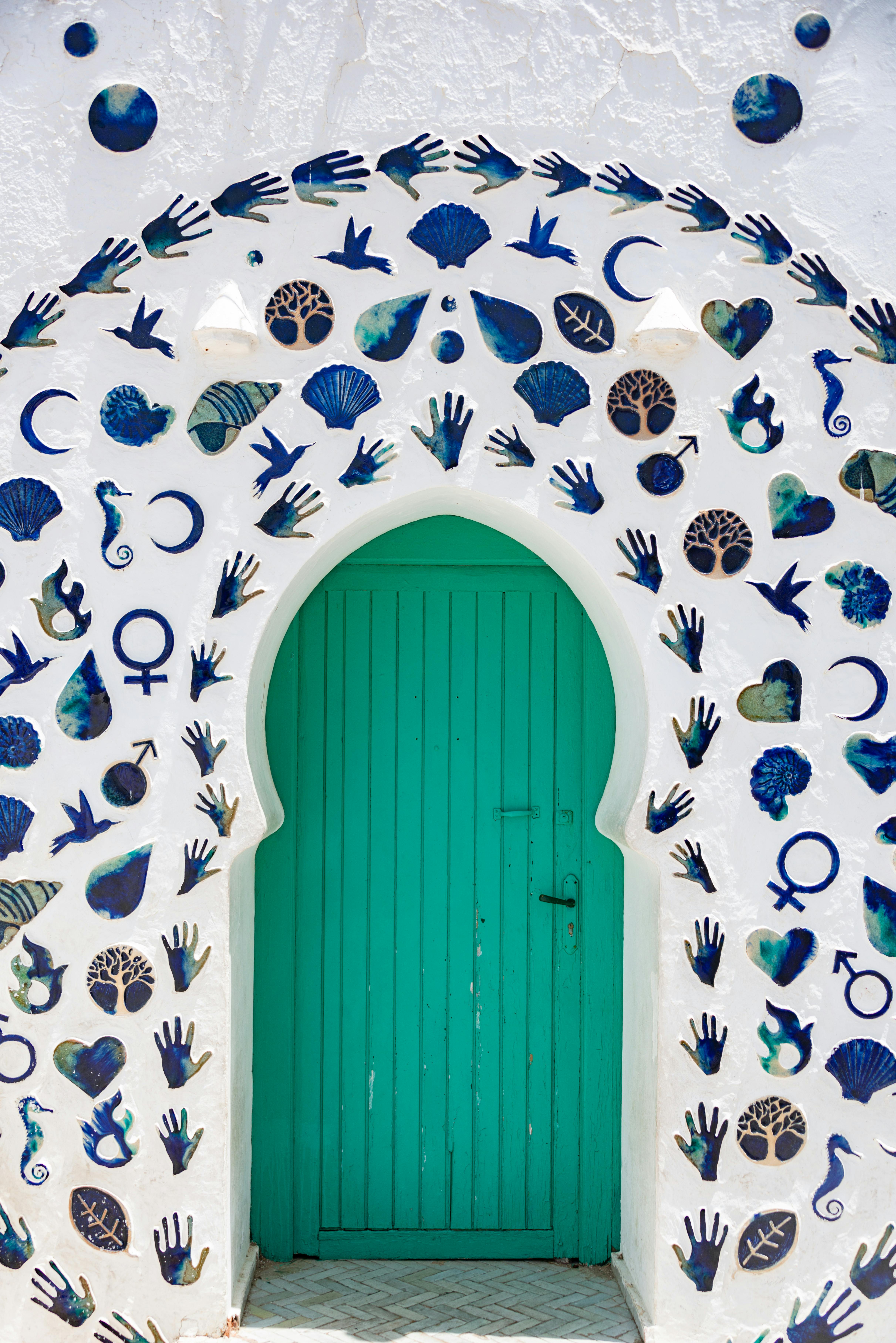 White wall with a teal door, adorned by a colorful ceramic arch featuring diverse symbols and motifs.