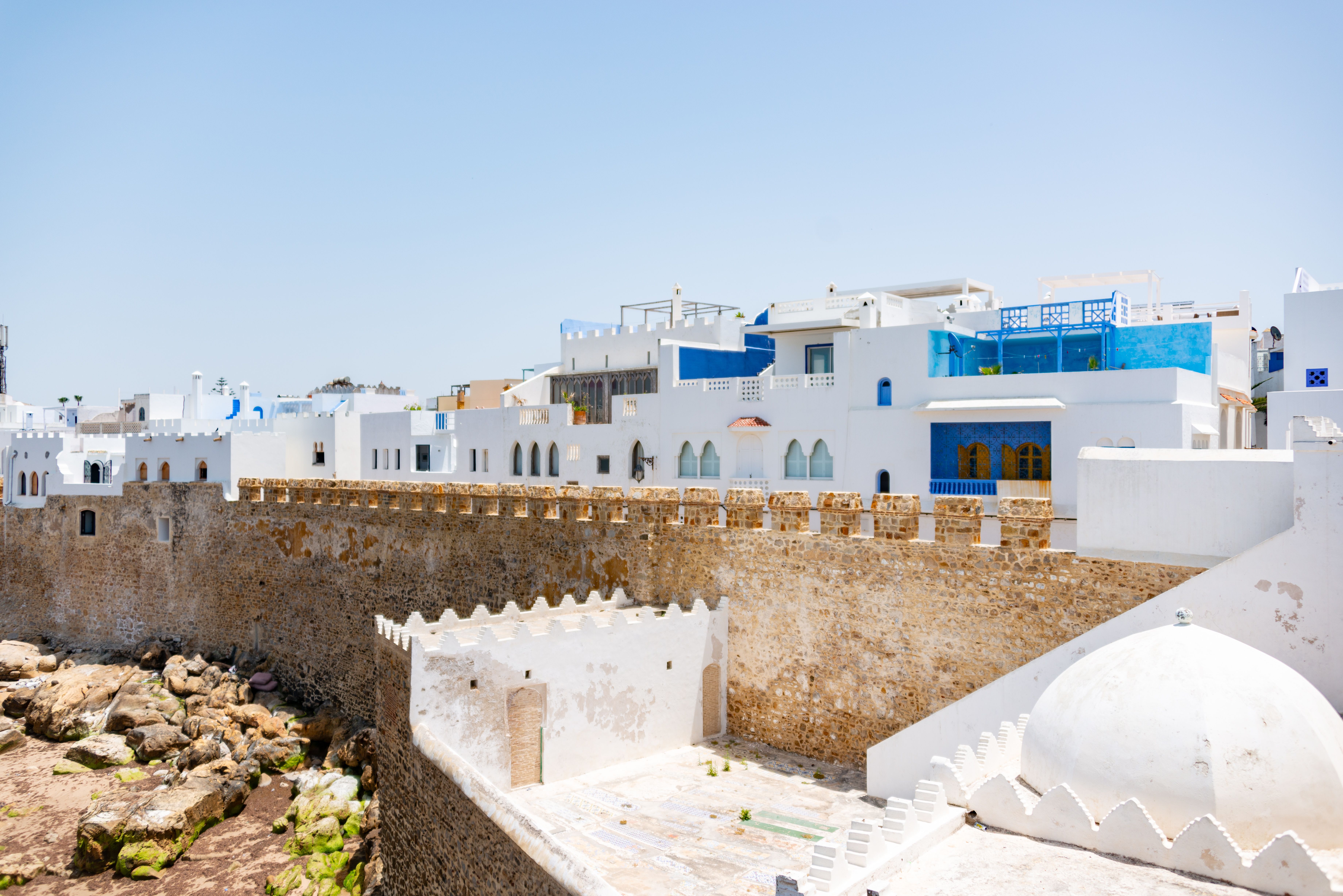 Scenic view of ancient city with white and blue houses and fortress walls under a clear blue sky. - Asilah