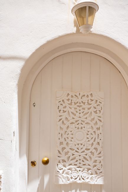 An ornate white arched door with a detailed decorative panel and brass knob, bathed in sunlight.