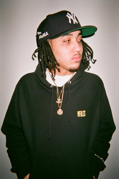 Photo of Man in Black Hoodie and Black New York Yankees Cap Posing In Front of White Background Looking Away