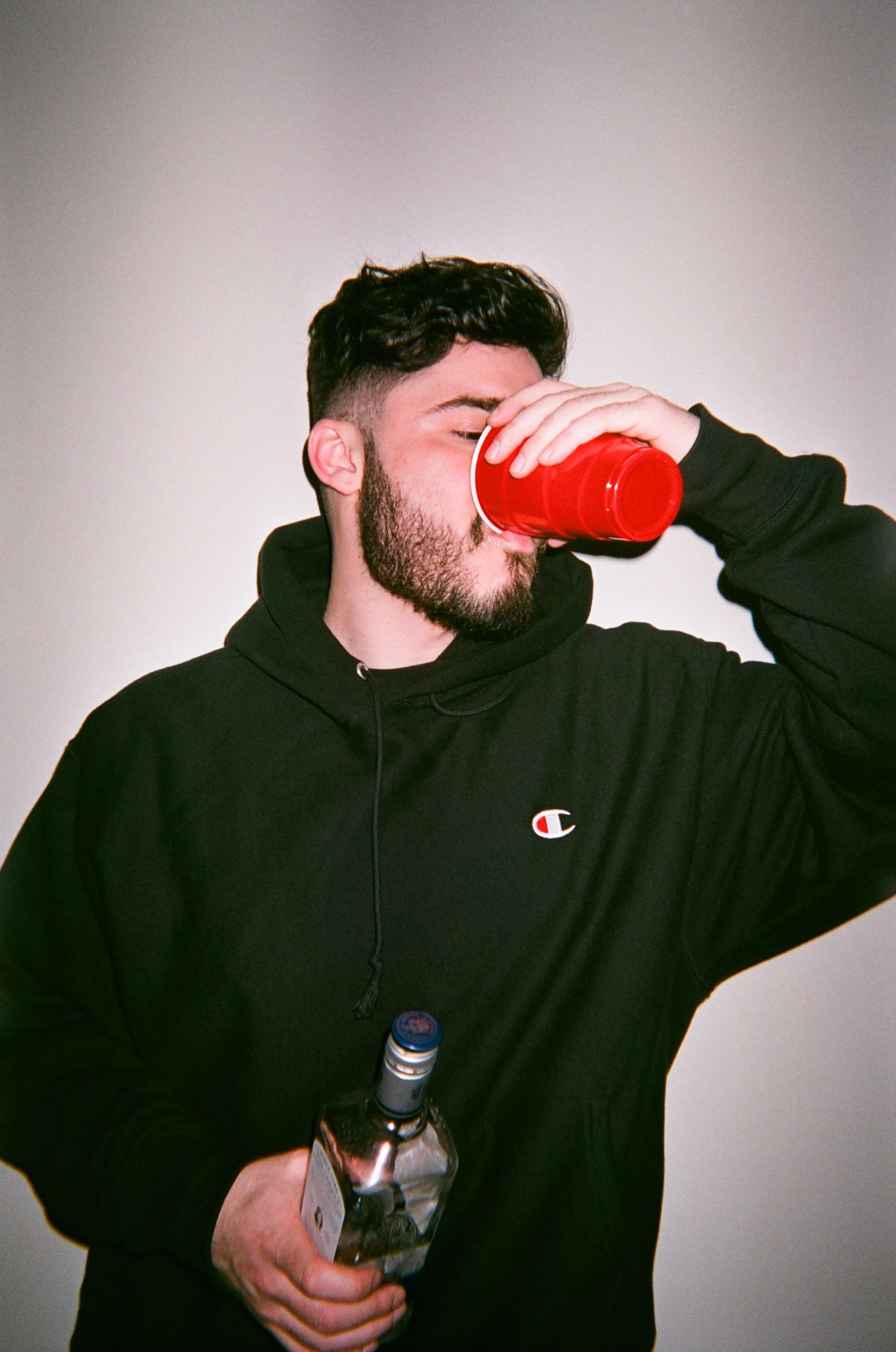 A bearded man in a black hoodie drinks from a red cup while holding a clear bottle.