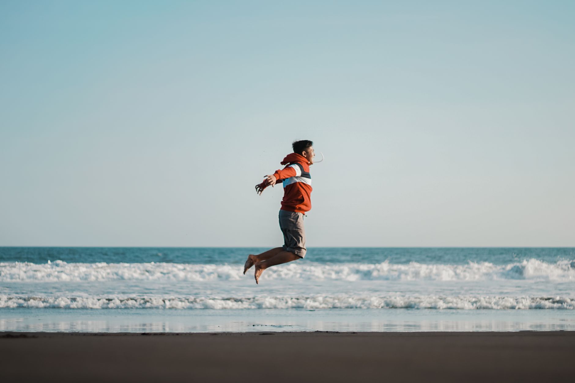 Man in red jacket jumping on the beach by the ocean, enjoying a carefree day outdoors.