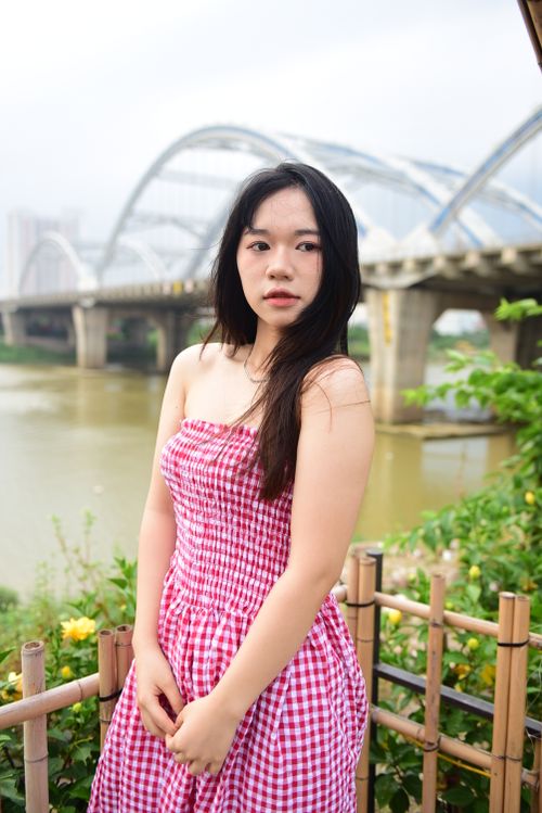 Free A woman in a strapless red gingham dress poses by a scenic riverside bridge. Stock Photo