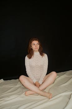 在工作室里，一位身着蕾丝、神情深思的女人盘腿坐在深色背景的布料上