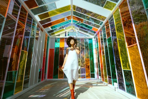 Photo of Smiling Woman in White Dress and Brown Boots Posing in Multicolored Glass House