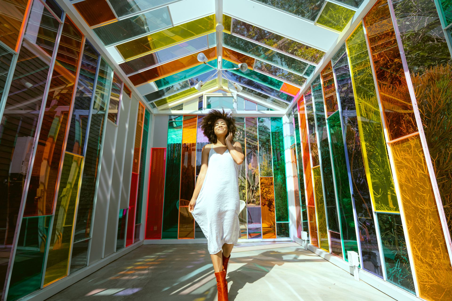 Photo of Smiling Woman in White Dress and Brown Boots Posing in Multicolored Glass House