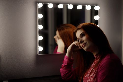 Free A woman in a red blouse gazes thoughtfully into a mirror surrounded by lights. Stock Photo