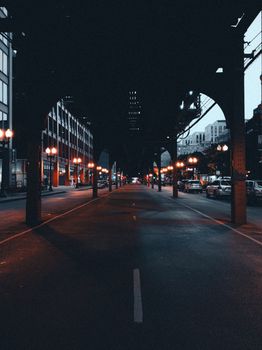 A dramatic view of an urban street under a viaduct, illuminated by street lamps at dusk.